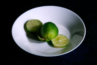 A white ceramic plate holds two lime halves and one whole lime. The limes have a vibrant green color and are positioned on a dark surface, contrasting with the bright white of the plate.
