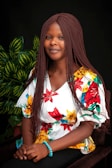 A woman with braided hair is sitting in front of a dark background, wearing a white blouse adorned with vibrant red, yellow, and green floral patterns. She has a serene expression, and a bright blue beaded bracelet adorns her wrist. There are green leafy plants in the background.