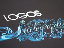 Elegant typography featuring the word 'LOGOS' in white capital letters, paired with an ornate script of 'Steelography' in a flowing, decorative blue font against a dark background.