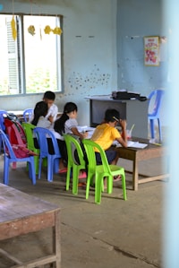 Several children are gathered around a low wooden table in a classroom, focusing on their studies. The room has simple furnishings with colorful plastic chairs, primarily green and blue, and a few educational materials visible. There are signs of wall wear, with paint peeling and chipped areas, and a window on one side letting in natural light. Decorations made of paper hang from the ceiling, adding a touch of color.