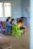 Several children are gathered around a low wooden table in a classroom, focusing on their studies. The room has simple furnishings with colorful plastic chairs, primarily green and blue, and a few educational materials visible. There are signs of wall wear, with paint peeling and chipped areas, and a window on one side letting in natural light. Decorations made of paper hang from the ceiling, adding a touch of color.