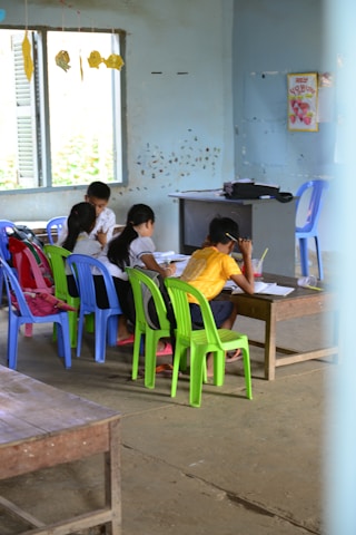 Several children are gathered around a low wooden table in a classroom, focusing on their studies. The room has simple furnishings with colorful plastic chairs, primarily green and blue, and a few educational materials visible. There are signs of wall wear, with paint peeling and chipped areas, and a window on one side letting in natural light. Decorations made of paper hang from the ceiling, adding a touch of color.