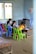 Several children are gathered around a low wooden table in a classroom, focusing on their studies. The room has simple furnishings with colorful plastic chairs, primarily green and blue, and a few educational materials visible. There are signs of wall wear, with paint peeling and chipped areas, and a window on one side letting in natural light. Decorations made of paper hang from the ceiling, adding a touch of color.
