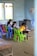 Several children are gathered around a low wooden table in a classroom, focusing on their studies. The room has simple furnishings with colorful plastic chairs, primarily green and blue, and a few educational materials visible. There are signs of wall wear, with paint peeling and chipped areas, and a window on one side letting in natural light. Decorations made of paper hang from the ceiling, adding a touch of color.