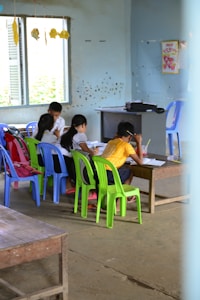 Several children are gathered around a low wooden table in a classroom, focusing on their studies. The room has simple furnishings with colorful plastic chairs, primarily green and blue, and a few educational materials visible. There are signs of wall wear, with paint peeling and chipped areas, and a window on one side letting in natural light. Decorations made of paper hang from the ceiling, adding a touch of color.