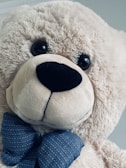A large, fluffy teddy bear with soft, light beige fur. It has dark, round eyes and a prominent black nose. The bear is adorned with a blue bow tie featuring a subtle pattern.