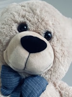 A large, fluffy teddy bear with soft, light beige fur. It has dark, round eyes and a prominent black nose. The bear is adorned with a blue bow tie featuring a subtle pattern.