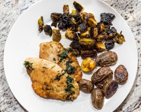 A white plate on a granite surface holds two pieces of pan-seared chicken breast garnished with herbs. Next to the chicken are roasted Brussels sprouts and a variety of small roasted potatoes, some with a golden, crispy exterior.