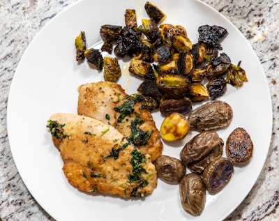 A white plate on a granite surface holds two pieces of pan-seared chicken breast garnished with herbs. Next to the chicken are roasted Brussels sprouts and a variety of small roasted potatoes, some with a golden, crispy exterior.