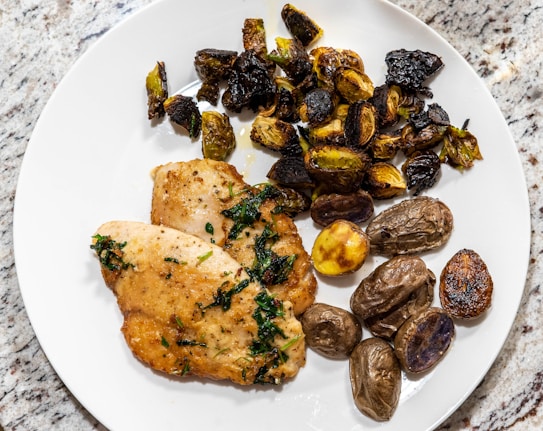 A white plate on a granite surface holds two pieces of pan-seared chicken breast garnished with herbs. Next to the chicken are roasted Brussels sprouts and a variety of small roasted potatoes, some with a golden, crispy exterior.
