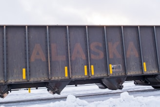 A large freight train car with the word 'ALASKA' prominently displayed on its side. The car is dark and weathered, with yellow vertical markings acting as accents. Snow covers the ground, suggesting a cold, wintry environment.