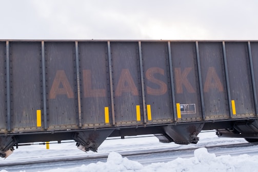 A large freight train car with the word 'ALASKA' prominently displayed on its side. The car is dark and weathered, with yellow vertical markings acting as accents. Snow covers the ground, suggesting a cold, wintry environment.