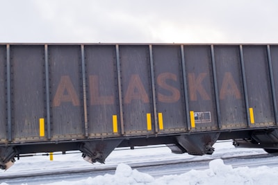 A large freight train car with the word 'ALASKA' prominently displayed on its side. The car is dark and weathered, with yellow vertical markings acting as accents. Snow covers the ground, suggesting a cold, wintry environment.