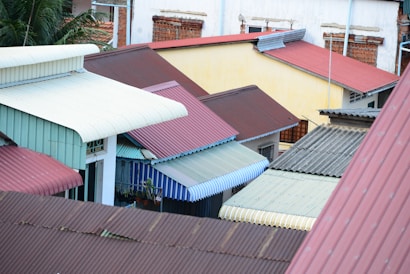 Multiple corrugated metal rooftops in various colors such as red, green, blue, and white are tightly packed together in an urban setting. Background trees and walls with small windows are visible.