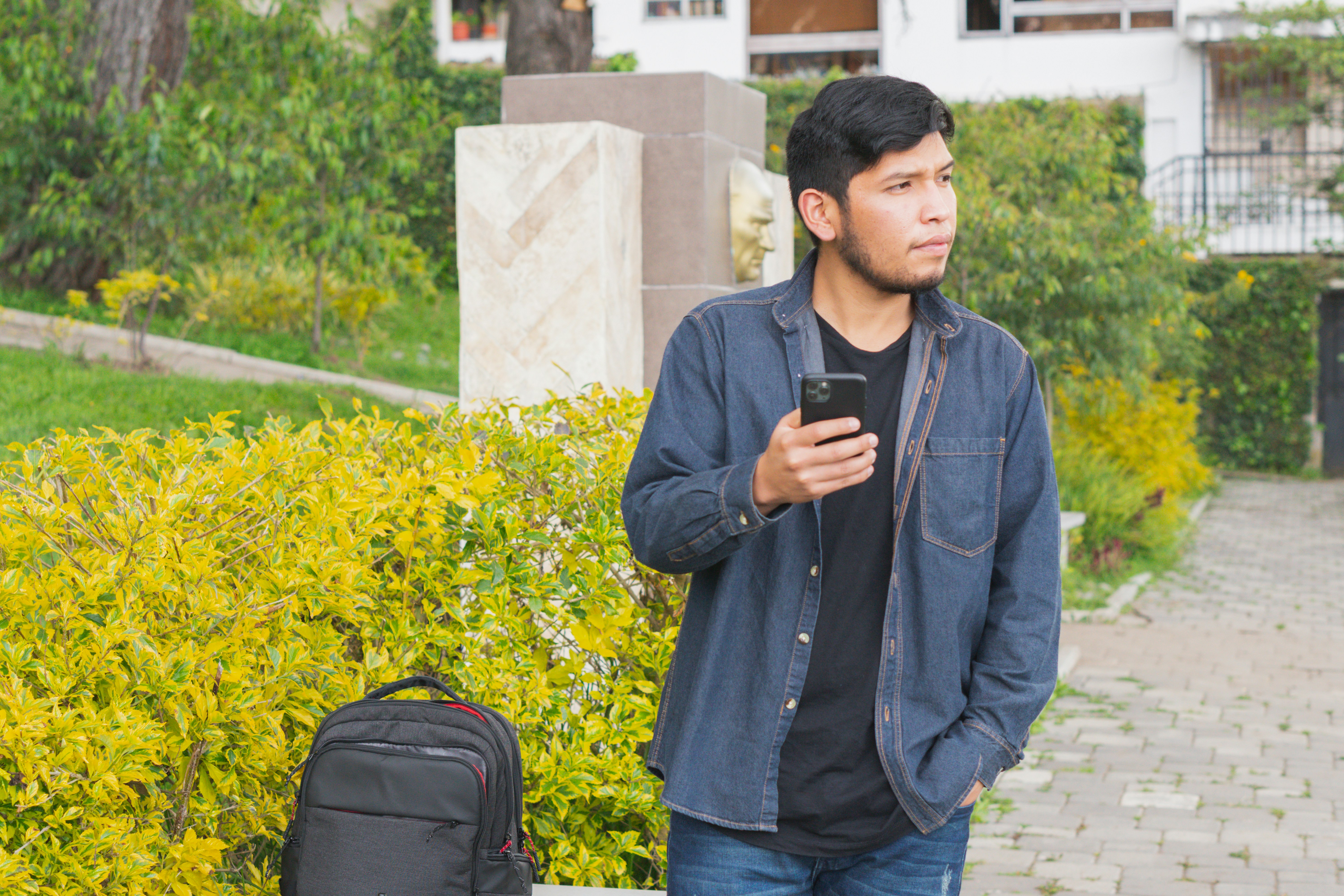 man in black jacket and blue denim jeans standing and holding black smartphone