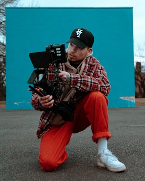 A man crouches on an asphalt surface holding a large camera with professional equipment attached. He wears a red and black checkered jacket, an embroidered baseball cap, red pants, and white sneakers. A teal wall forms the background, contrasting with the muted tones of the setting.