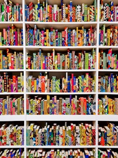 A series of bookshelves filled with books that have vividly patterned and colorful covers. The books are arranged neatly, creating a visually striking display of geometric and abstract designs. Each book seems to have a unique combination of colors and patterns, ranging from stripes and dots to floral and geometric motifs.