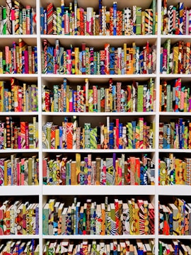 A series of bookshelves filled with books that have vividly patterned and colorful covers. The books are arranged neatly, creating a visually striking display of geometric and abstract designs. Each book seems to have a unique combination of colors and patterns, ranging from stripes and dots to floral and geometric motifs.