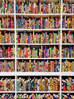 A series of bookshelves filled with books that have vividly patterned and colorful covers. The books are arranged neatly, creating a visually striking display of geometric and abstract designs. Each book seems to have a unique combination of colors and patterns, ranging from stripes and dots to floral and geometric motifs.