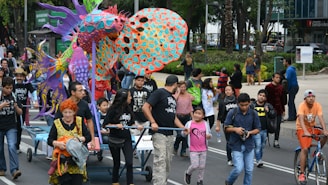 A lively parade features a colorful, large alebrije, a fantastical creature made of various vibrant materials. People of different ages, including children and adults, participate by pushing the structure. One person rides a bicycle, and others walk alongside, capturing the scene with cameras. The street is bustling with activity, and a crowd is gathered to watch.
