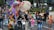 A lively parade features a colorful, large alebrije, a fantastical creature made of various vibrant materials. People of different ages, including children and adults, participate by pushing the structure. One person rides a bicycle, and others walk alongside, capturing the scene with cameras. The street is bustling with activity, and a crowd is gathered to watch.