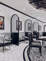 An elegant indoor dining area features modern decor with a blend of classic and contemporary design. The room has tiled floors with intricate patterns and high-backed chairs surrounding round tables. Decorative wall dividers and framed artwork contribute to the sophisticated ambiance. Subtle lighting fixtures hang from the ceiling, contributing to a sleek and stylish atmosphere.