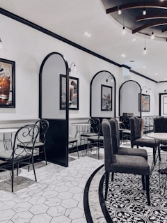 An elegant indoor dining area features modern decor with a blend of classic and contemporary design. The room has tiled floors with intricate patterns and high-backed chairs surrounding round tables. Decorative wall dividers and framed artwork contribute to the sophisticated ambiance. Subtle lighting fixtures hang from the ceiling, contributing to a sleek and stylish atmosphere.