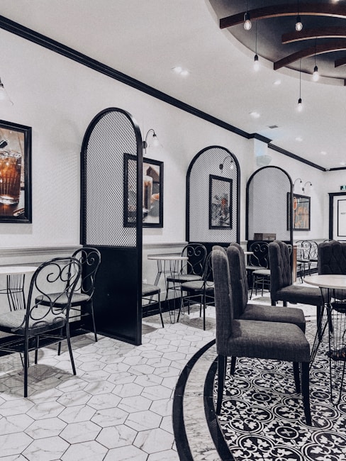 An elegant indoor dining area features modern decor with a blend of classic and contemporary design. The room has tiled floors with intricate patterns and high-backed chairs surrounding round tables. Decorative wall dividers and framed artwork contribute to the sophisticated ambiance. Subtle lighting fixtures hang from the ceiling, contributing to a sleek and stylish atmosphere.