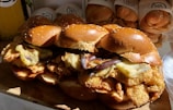 Four large sandwiches with sesame seed buns, stacked with crispy fried chicken and grilled eggplant slices. The sandwiches are arranged on a wooden surface, with a drink in the background.