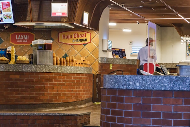 The scene depicts a chaat counter inside what appears to be a food establishment. There is a brick and stone counter with stacks of small bowls and various containers likely holding ingredients or sauces. The background wall has two signs indicating 'Laxmi Lunch Home' and 'Raju Chaat Bhandar'. To the right, a person is partially visible behind a countertop with a glass partition, presumably a staff member.