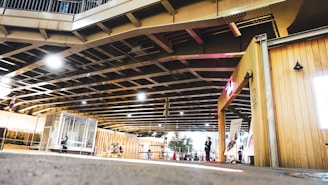 A modern urban space beneath a large bridge, featuring geometric structures and a small gathering of people. Light fixtures hang from the ceiling, illuminating a wooden kiosk with signs. The area is enclosed with railing, and small plants are visible nearby.