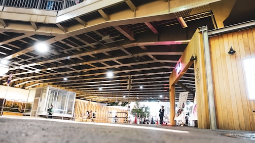 A modern urban space beneath a large bridge, featuring geometric structures and a small gathering of people. Light fixtures hang from the ceiling, illuminating a wooden kiosk with signs. The area is enclosed with railing, and small plants are visible nearby.