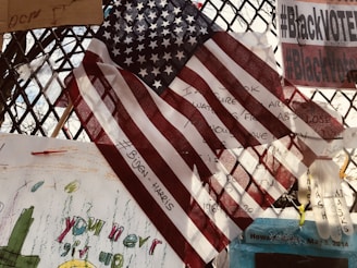 An American flag is prominently displayed, overlapping various notes and messages on a metal fence. The flag partially covers handwritten texts and a hashtag referencing political figures. Signs nearby include phrases on activism and encouragement with colorful drawings. Some objects are attached to the fence, including a plastic glove.
