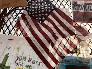 An American flag is prominently displayed, overlapping various notes and messages on a metal fence. The flag partially covers handwritten texts and a hashtag referencing political figures. Signs nearby include phrases on activism and encouragement with colorful drawings. Some objects are attached to the fence, including a plastic glove.