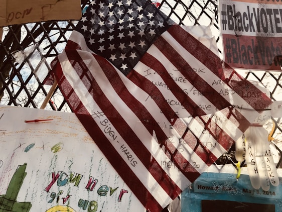 An American flag is prominently displayed, overlapping various notes and messages on a metal fence. The flag partially covers handwritten texts and a hashtag referencing political figures. Signs nearby include phrases on activism and encouragement with colorful drawings. Some objects are attached to the fence, including a plastic glove.
