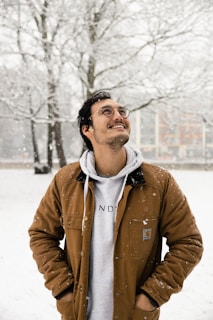 A smiling man buttoning up a thick brown parka while snow gently falls around him.
