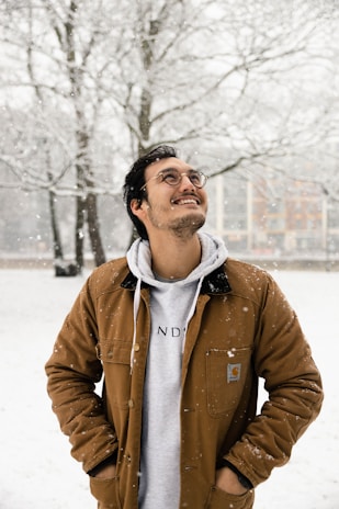 A smiling man buttoning up a thick brown parka while snow gently falls around him.