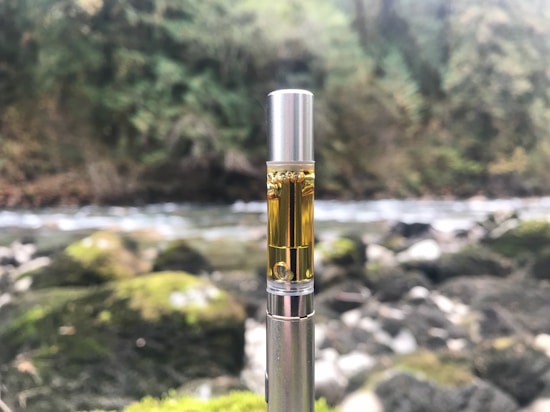 A vape pen cartridge is pictured with a natural background of a flowing river surrounded by green foliage and rocks. The image is focused on the cartridge, showcasing its metallic and transparent structure containing a golden liquid.