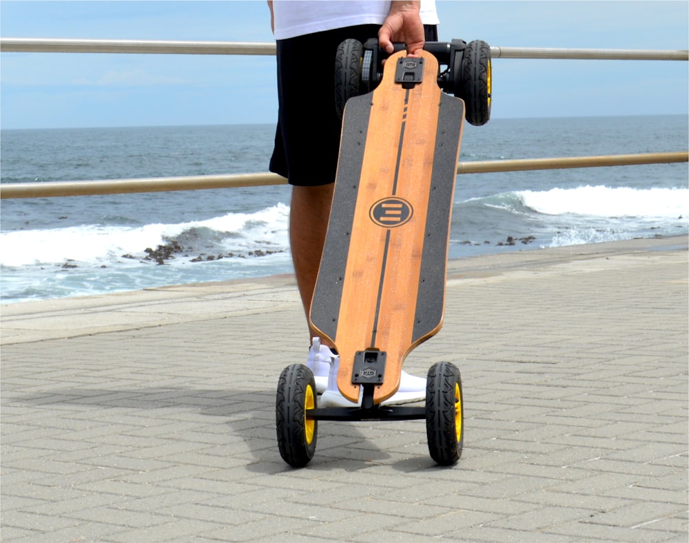 Persona en pantalones cortos negros y camisa blanca sosteniendo una patineta de madera marrón en la playa durante el día