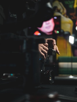 A person is adjusting a camera on a stabilizer in a dimly lit environment, with a blurred background featuring colorful shapes and lights. The focus is on the hands and equipment.