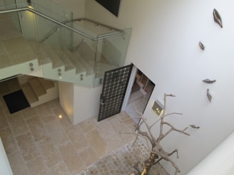 A modern interior featuring a staircase with glass railings and stone tile flooring. A decorative metallic sculpture resembling a tree is prominently displayed near a wall adorned with abstract metallic leaves. Subdued lighting creates a soft ambiance.