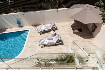 A serene outdoor space featuring a swimming pool with clear blue water and wooden decking. Two striped chaise lounges with white towels are positioned beside a small round table. A large beige umbrella provides shade to part of the area. Glass barriers and a lush green plant enhance the modern aesthetic, while a white wall and railing enclose the area.