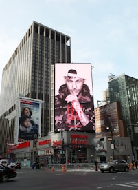 A bustling urban scene features large buildings and digital billboards. The central billboard displays a person wearing a hoodie and cap, with a finger pressed to their lips. Below, the street is alive with vehicles and pedestrians, indicating a busy city area. Additional billboards promote various products and brands.