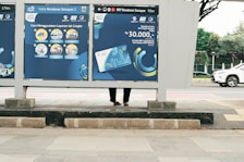 A public transportation sign at a bus stop displays information about using a transit service. The sign features blue and yellow illustrations and text with instructions and pricing. Behind the sign, part of a person's legs and feet are visible as they stand on the sidewalk. In the background, a white car drives down the street, and a tree-lined area can be seen.