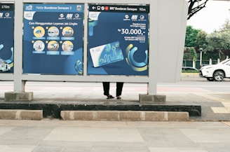 A public transportation sign at a bus stop displays information about using a transit service. The sign features blue and yellow illustrations and text with instructions and pricing. Behind the sign, part of a person's legs and feet are visible as they stand on the sidewalk. In the background, a white car drives down the street, and a tree-lined area can be seen.