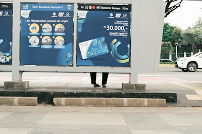 A public transportation sign at a bus stop displays information about using a transit service. The sign features blue and yellow illustrations and text with instructions and pricing. Behind the sign, part of a person's legs and feet are visible as they stand on the sidewalk. In the background, a white car drives down the street, and a tree-lined area can be seen.