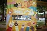 A colorful mural features various handprints in different colors and styles. The central themes include messages of love and kindness. Phrases like 'Love and Kindness are Never Wasted', 'they always make a difference', and 'Bless the receiver, bless the giver' are incorporated into the design. The mural is situated outdoors near a park with trees and people walking by.