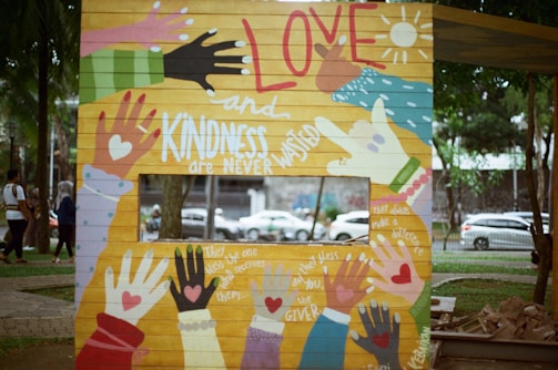 A colorful mural features various handprints in different colors and styles. The central themes include messages of love and kindness. Phrases like 'Love and Kindness are Never Wasted', 'they always make a difference', and 'Bless the receiver, bless the giver' are incorporated into the design. The mural is situated outdoors near a park with trees and people walking by.