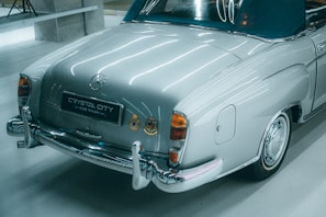 A classic car with a silver body and shiny chrome details is parked indoors. It has a dark blue convertible roof and vintage branding on the rear. The environment is well-lit with sleek, modern lighting that reflects off the polished surfaces.