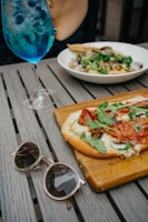 A table setting with a wooden surface holds a flatbread topped with melted cheese, arugula, and slices of cured meat. Next to it are stylish sunglasses. A vibrant blue cocktail with fruit floats nearby, and in the background, a bowl of pasta with clams and greens is partially visible.